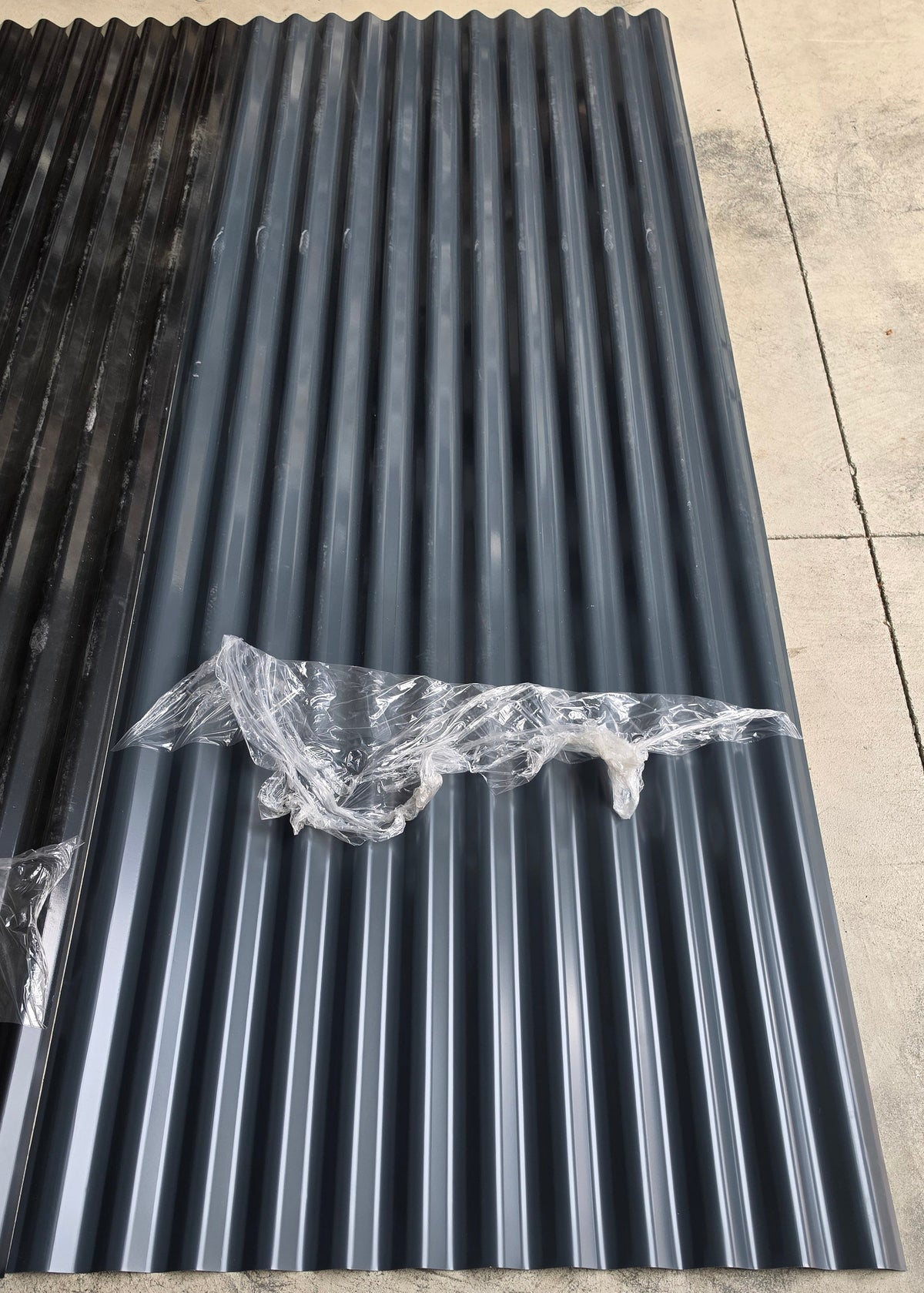 A long sheet of dark gray corrugated plastic or metal roofing panels laid on a concrete floor, with a clear protective cover section exposed near the center.