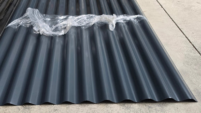 Black corrugated roofing panels with a matte finish, showing protective clear cover wrap, laid out on a concrete floor.