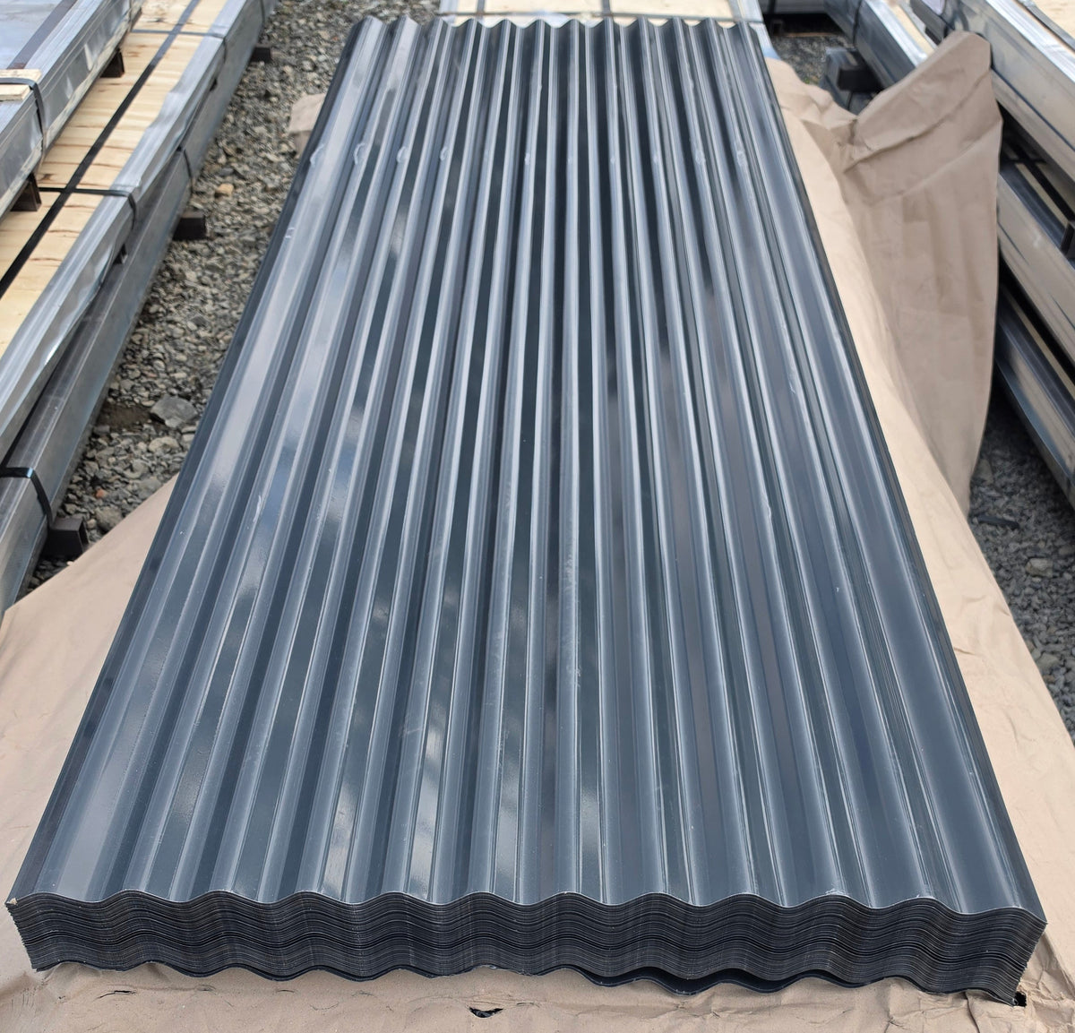 A long rectangular dark gray corrugated metal roofing sheet resting on brown paper, with other roofing materials stacked in the background on a gravel surface.