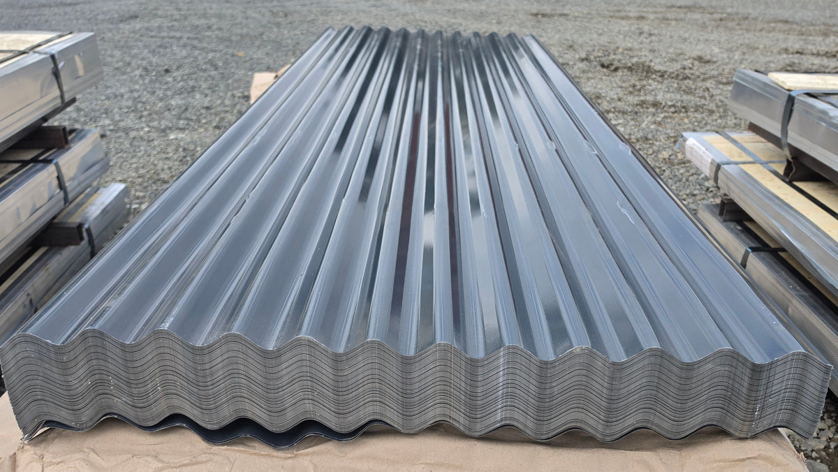 A stack of galvanized corrugated metal roofing panels resting on a tan sheet of paper on an outdoor gravel surface.