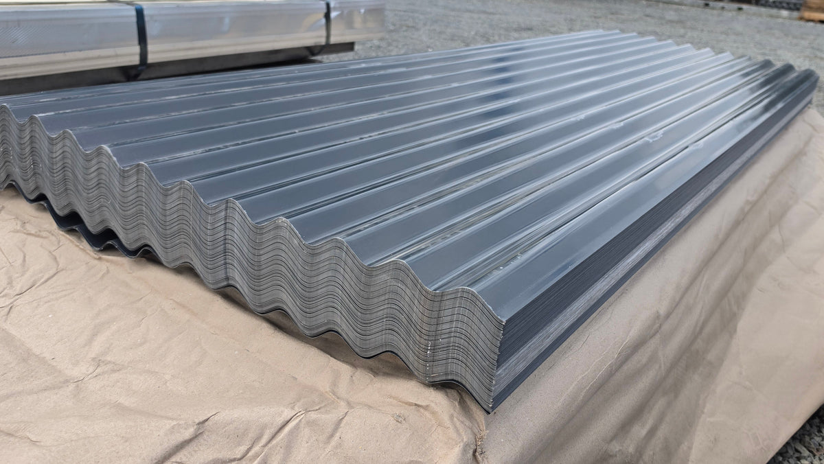 A large stack of galvanized corrugated metal roofing panels resting on a tan protective tarp in an outdoor warehouse or yard setting.