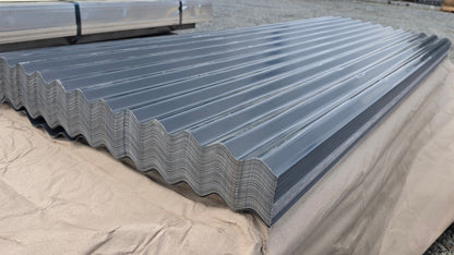 A large stack of galvanized corrugated metal roofing panels resting on a tan protective tarp in an outdoor warehouse or yard setting.