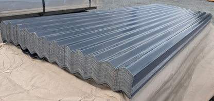 A large stack of galvanized corrugated metal roofing panels resting on a tan protective tarp in an outdoor warehouse or yard setting.
